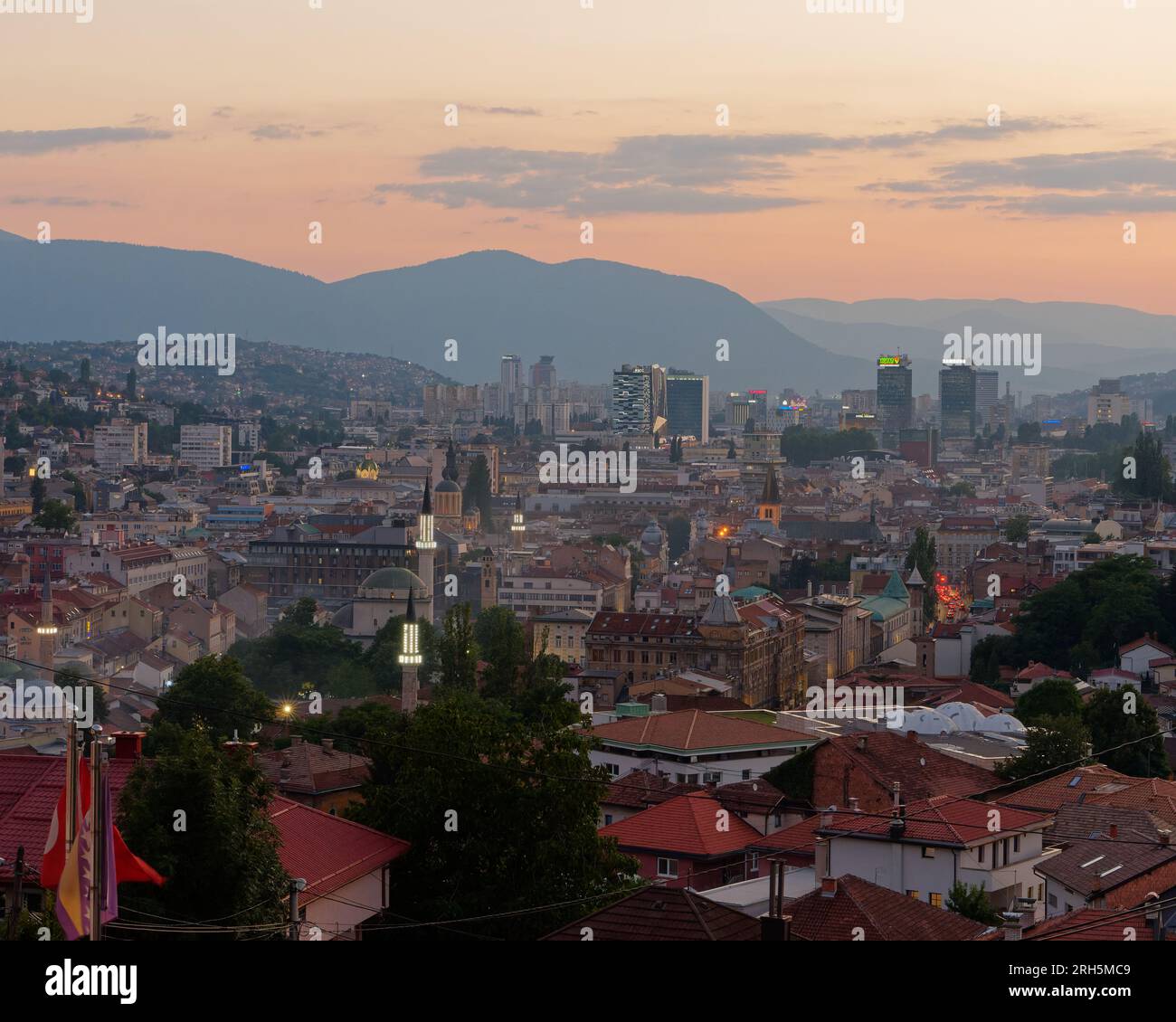 Vista sopraelevata su Sarajevo al tramonto. Minareti illuminati e condomini e montagne sullo sfondo, Bosnia ed Erzegovina, 13 agosto 2023. Foto Stock
