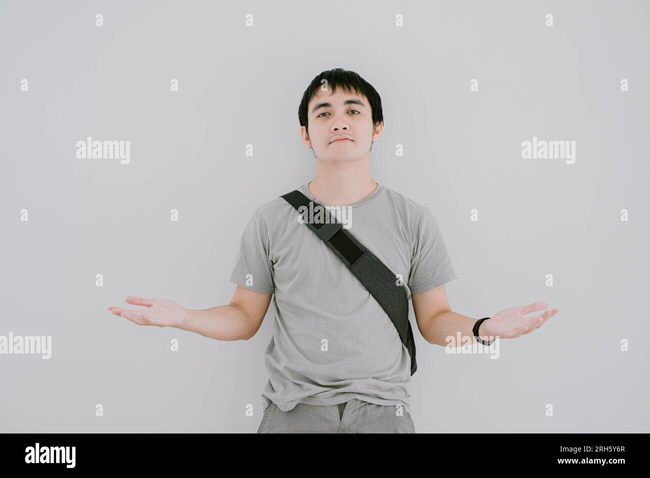 La messa a fuoco selettiva di un giovane asiatico che indossa una T-shirt casual verde salvia, uno smartwatch e una borsa a tracolla è un gesto confuso o non lo sa con le braccia sollevate Foto Stock