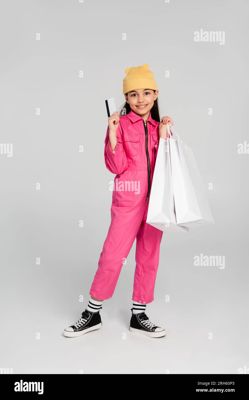 happy girl con berretto e outfit alla moda con carta di credito grigia, shopping per bambini, moda, gioia Foto Stock