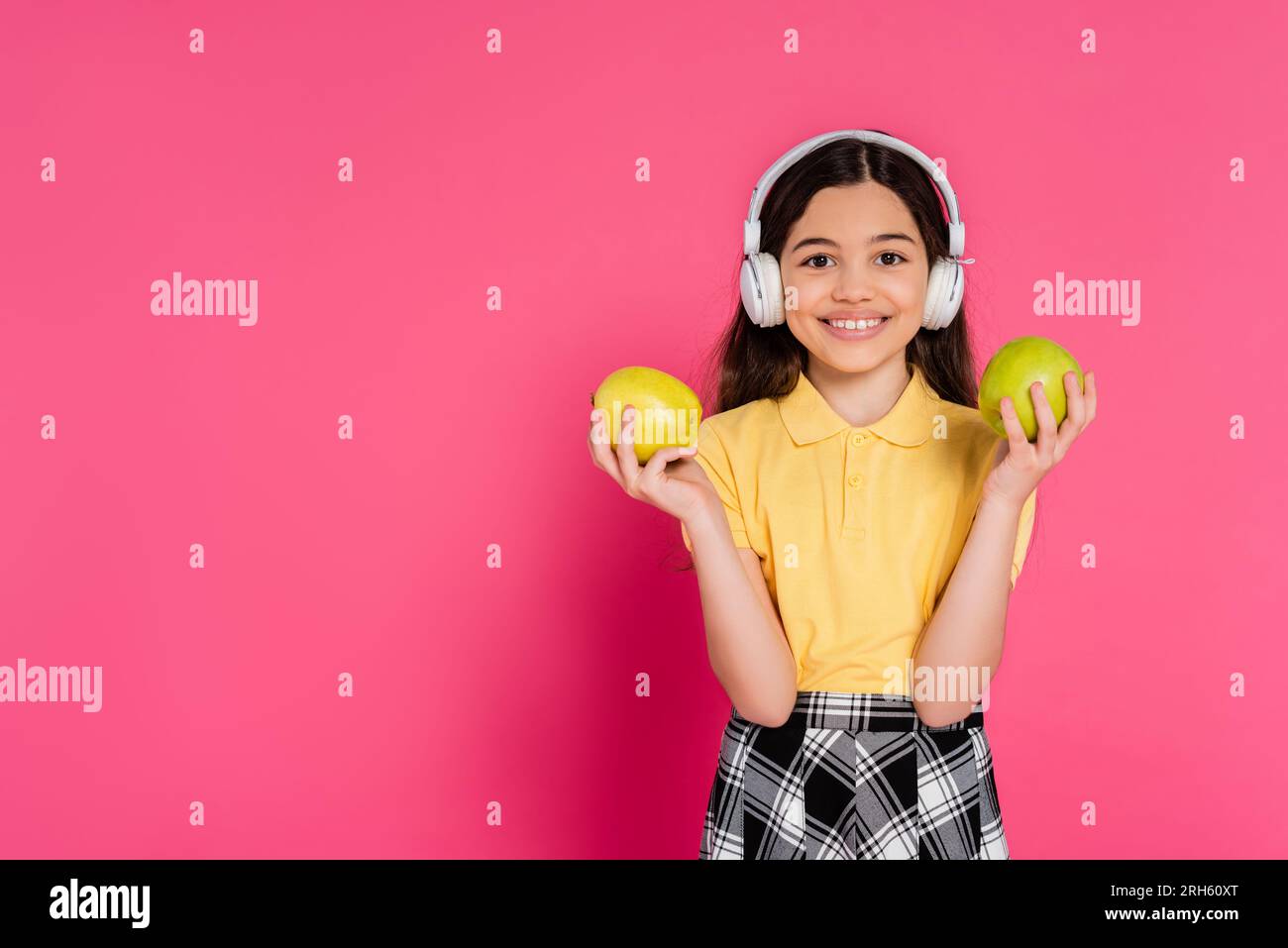 studentessa sorpresa con le cuffie wireless che reggono mela e zaino, sfondo rosa, studentessa Foto Stock