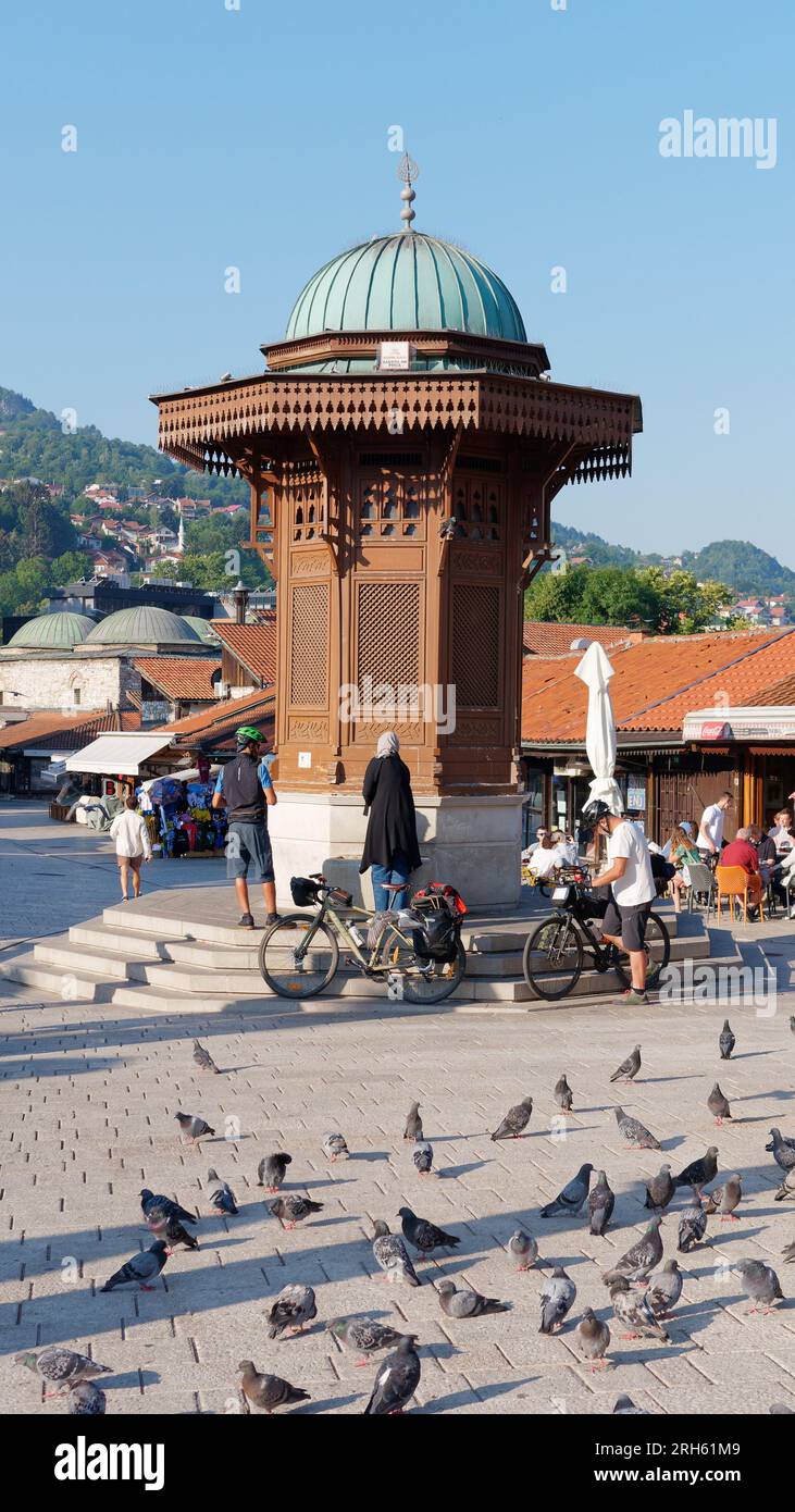 Ciclisti e piccioni vicino alla fontana Sebilj nel quartiere Baščaršija di Sarajevo, Bosnia ed Erzegovina, 14 agosto 2023. Foto Stock