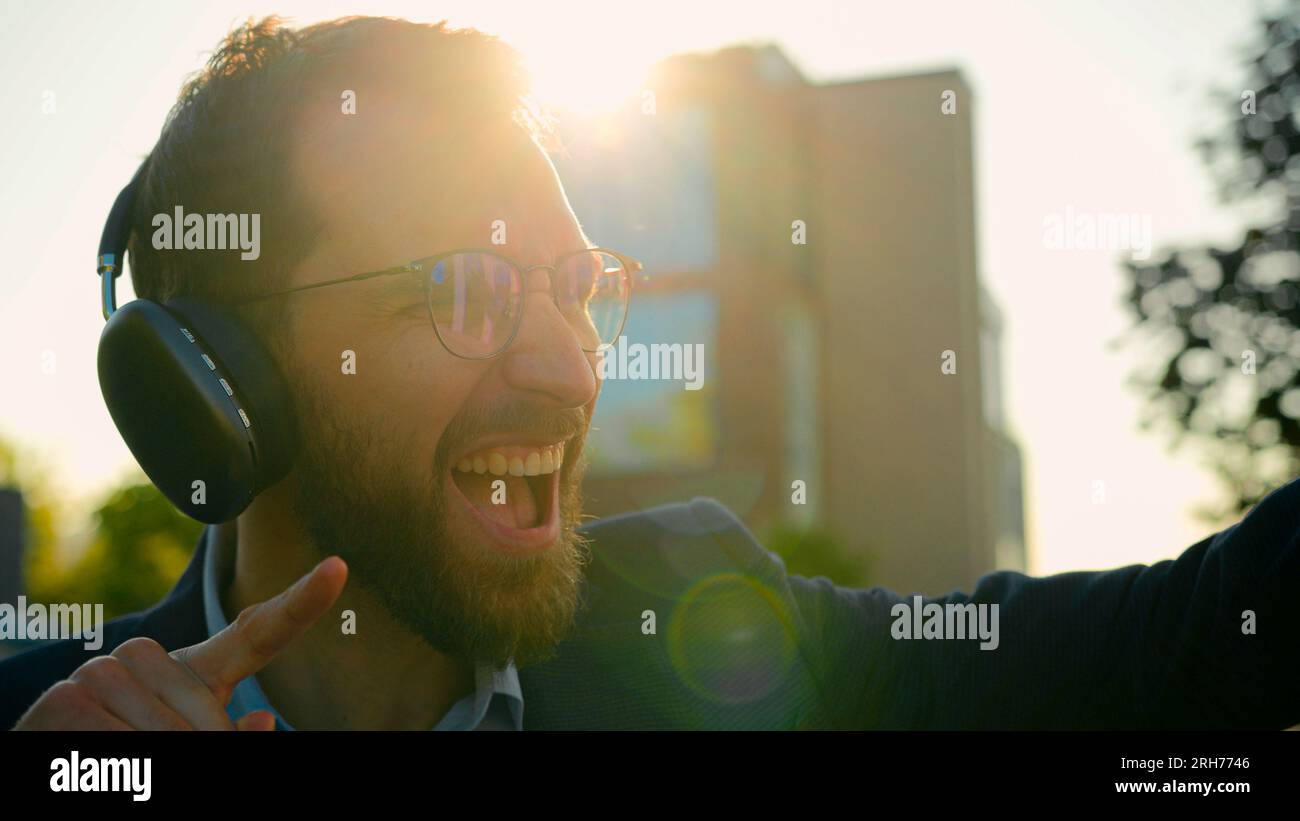 L'eccitato uomo d'affari caucasico ascolta la musica ballando con le cuffie all'aperto un uomo d'affari felice si diverte a cantare alla luce del sole della città Foto Stock