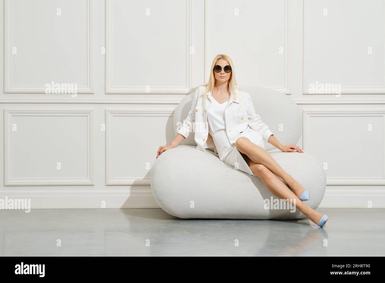 Donna sicura di sé, con giacca bianca e minigonna, si siede sul pouf in una stanza luminosa Foto Stock