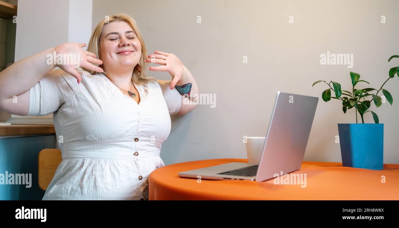 Una donna sorridente e sorridente seduta a un tavolo con un computer portatile, goditi la pausa caffè in un luogo di lavoro domestico. Foto Stock