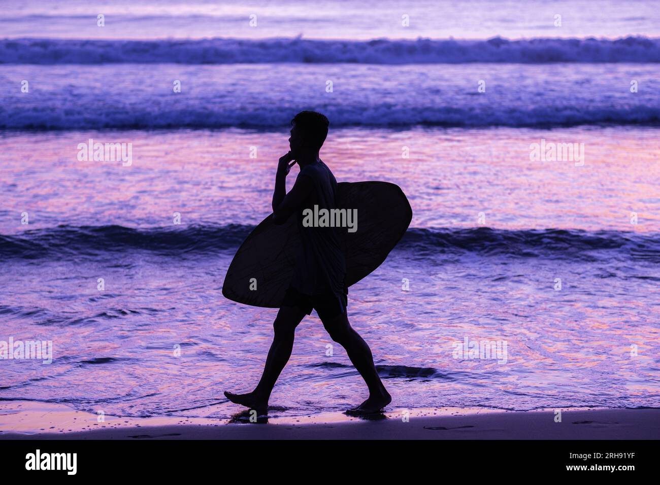 Silhouette di surfisti che portano la tavola da surf sulla spiaggia al tramonto con vibrazioni viola Foto Stock