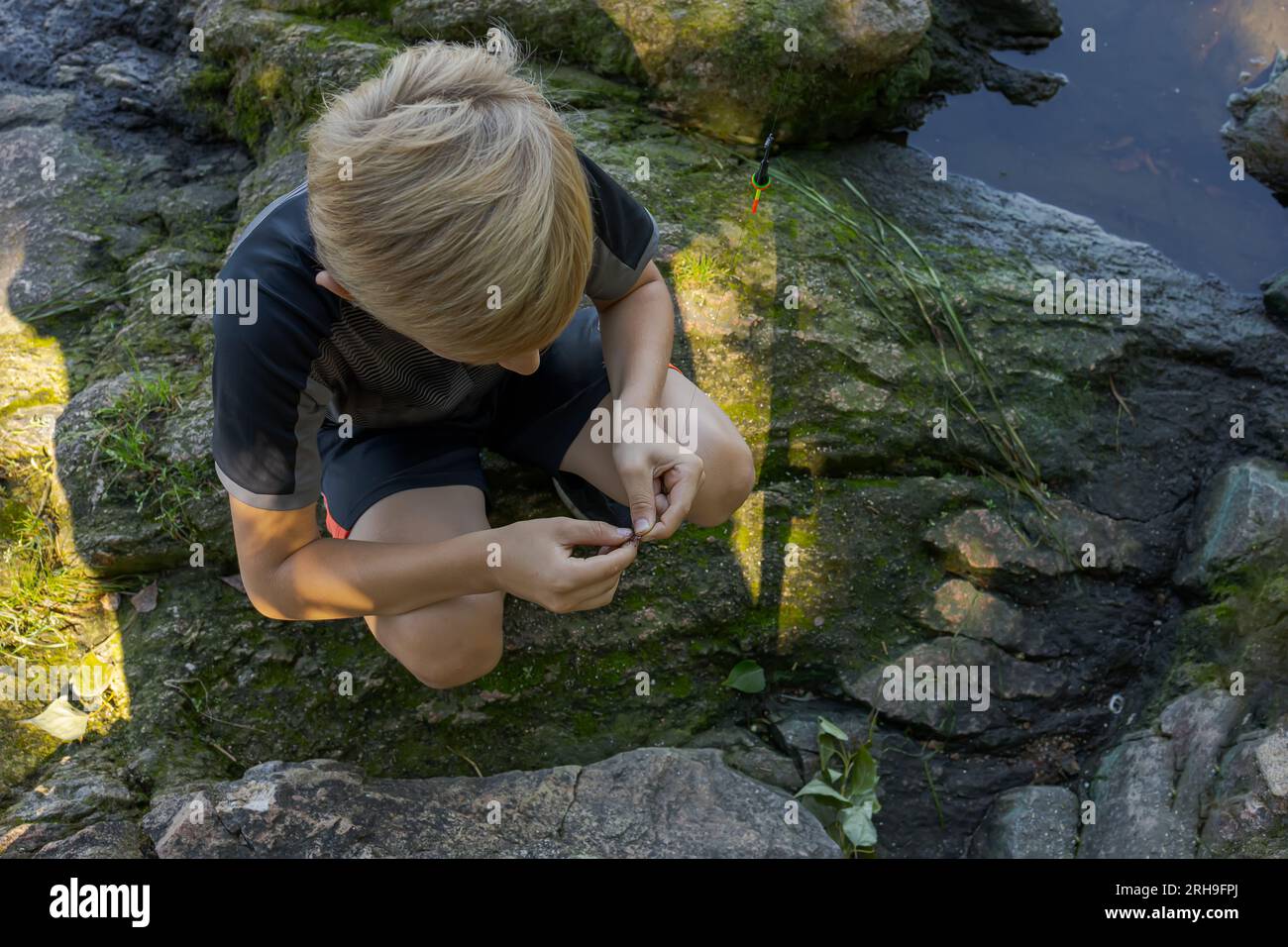 Un ragazzo seduto sulle rocce vicino al fiume equivale il gancio di un'esca. Pesca sportiva sul fiume in estate. Foto Stock
