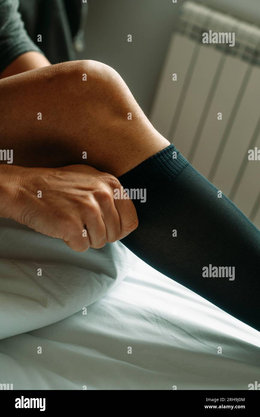 un uomo seduto sul letto indossa un calzino a compressione nero, con la luce del giorno Foto Stock