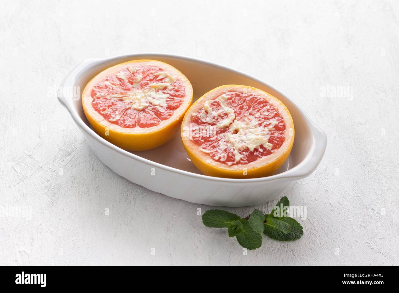 Piatto da forno in ceramica bianca metà di pompelmo rosso cosparso di burro su un tavolo grigio chiaro. Fase di cottura, cottura di un delizioso dessert. Foto Stock