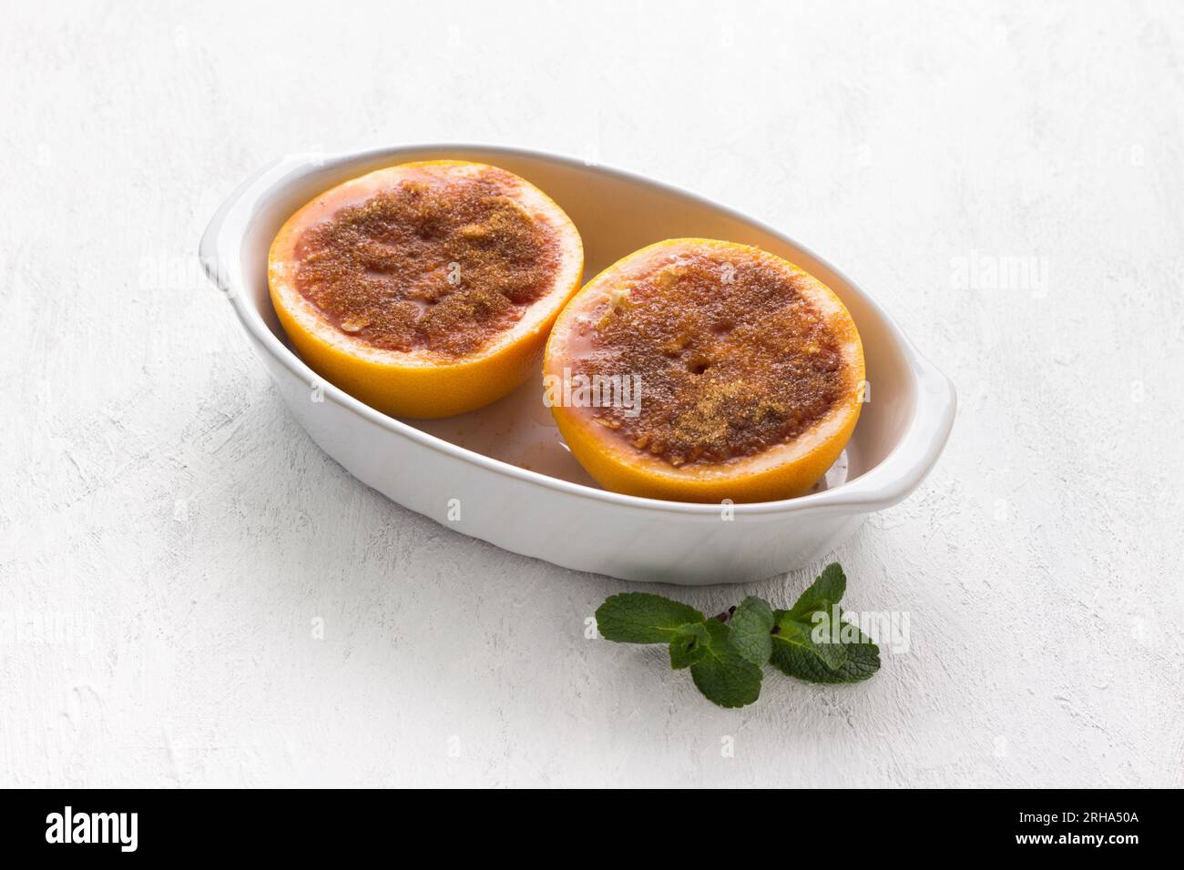 Piatto da forno in ceramica bianca con metà di pompelmo rosso cosparso di burro e cosparso di zucchero e cannella su un tavolo grigio chiaro. Fase di cottura, Co Foto Stock