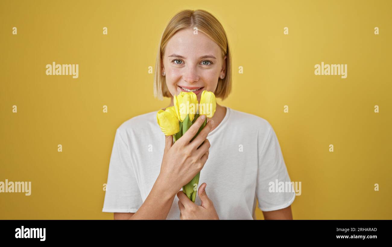 Giovane donna bionda che odora fiori sorridenti su sfondo giallo isolato Foto Stock