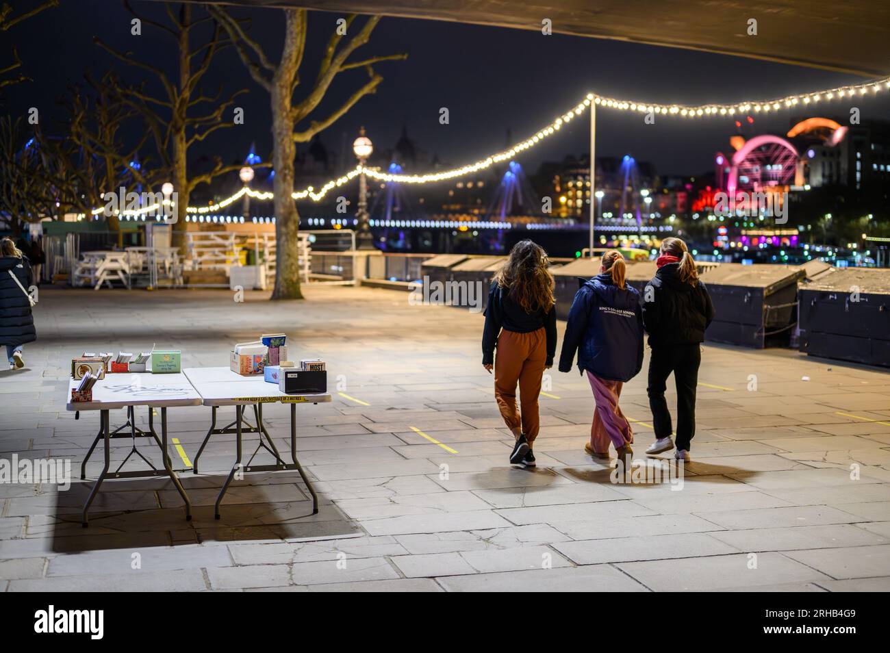 LONDRA - 22 aprile 2023: Passeggiata notturna: Le donne passano una bancarella su Southbank, tra le luci della città di Londra. La stazione ferroviaria di Charing Cross aggiunge un tocco di vivacità Foto Stock