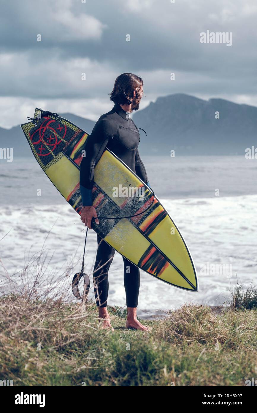 Corpo completo di un giovane atleta maschile con barba muscolare in muta nera con tavola da surf in mano in piedi sulla costa erbosa e guardando lontano dopo aver praticato su Foto Stock
