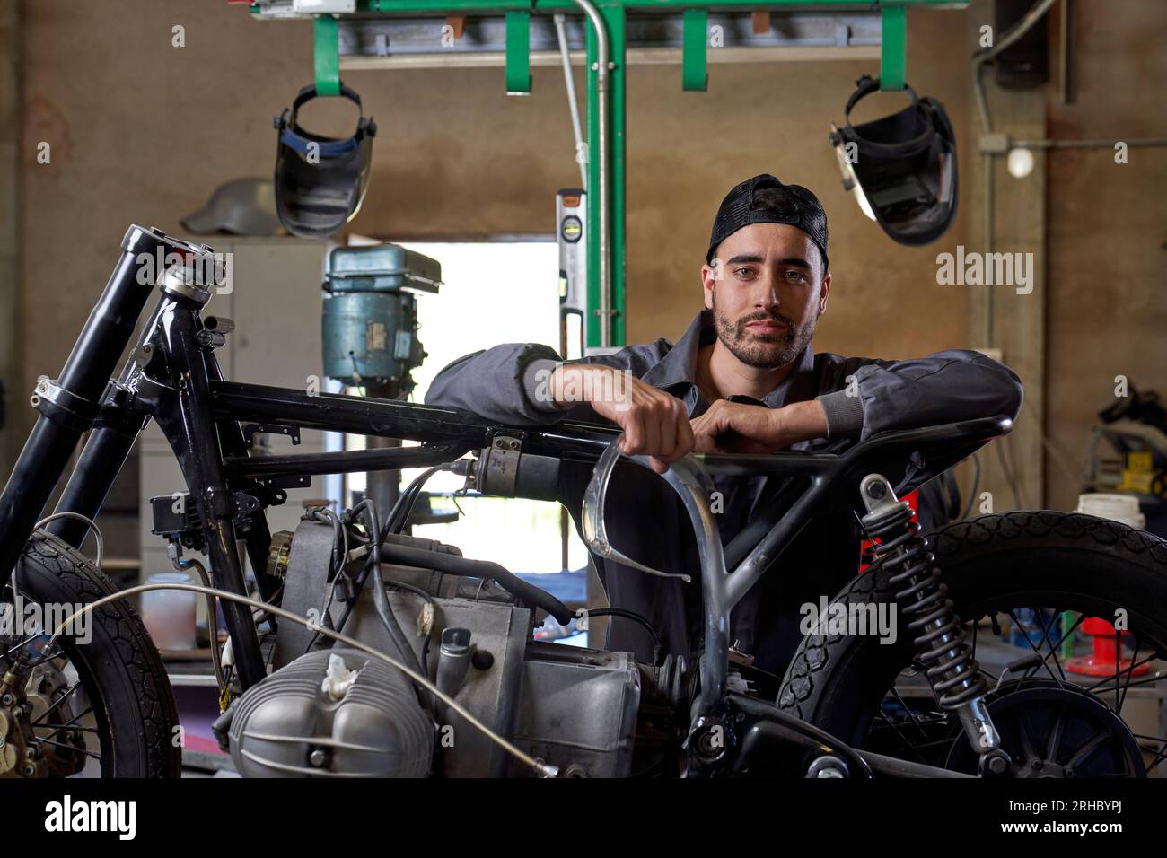 Tecnico maschio barbuto sicuro che indossa l'uniforme appoggiandosi alla moto in officina e guardando la fotocamera Foto Stock