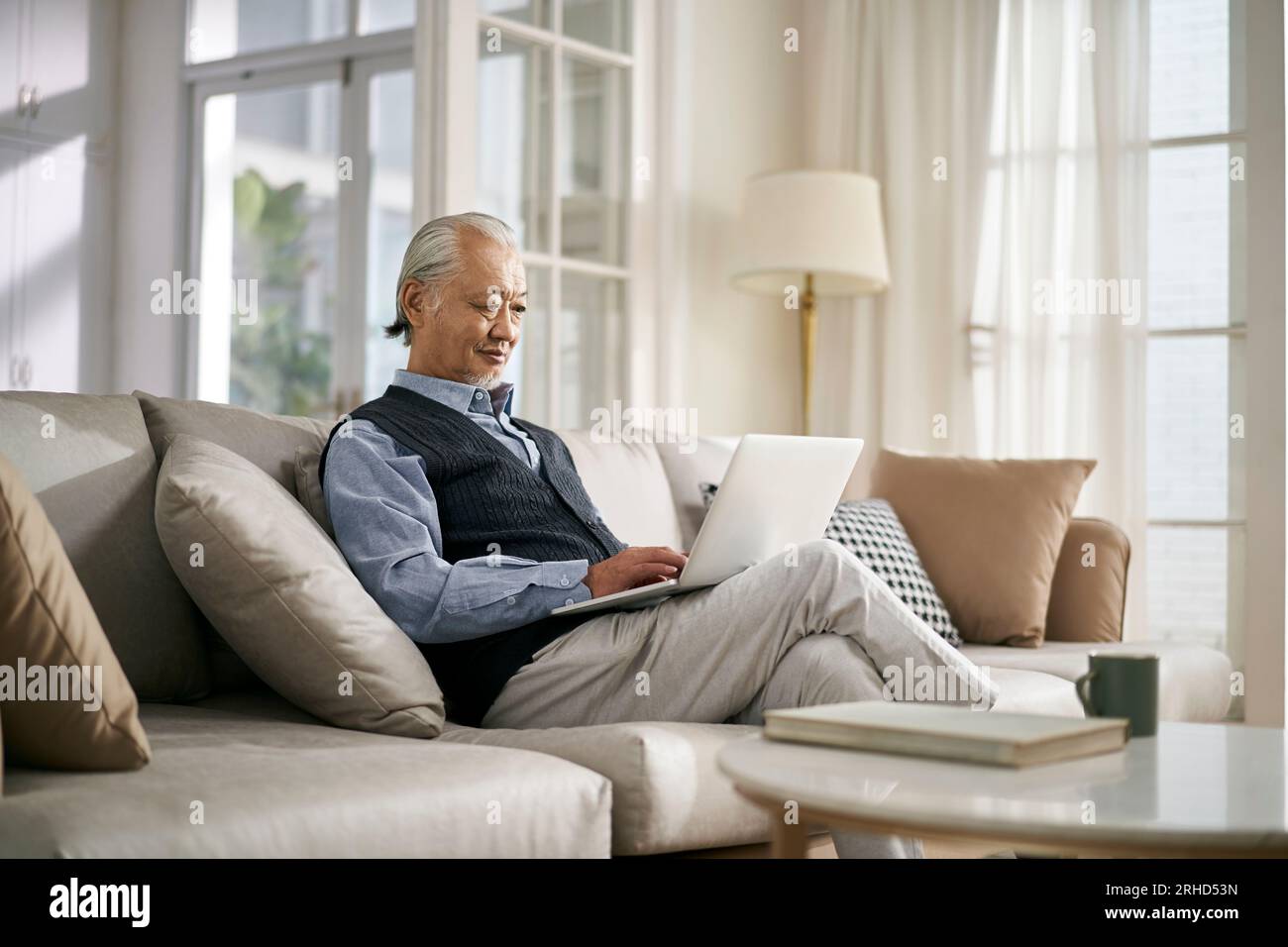 vista laterale di un anziano asiatico seduto sul divano a casa con un computer portatile Foto Stock