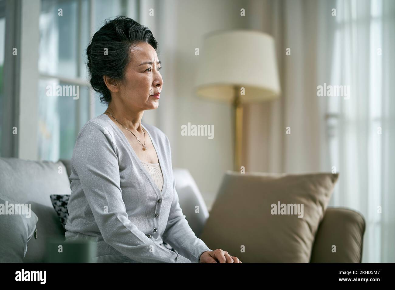 vista laterale di una donna asiatica anziana e triste seduta da sola sul divano in salotto a casa Foto Stock