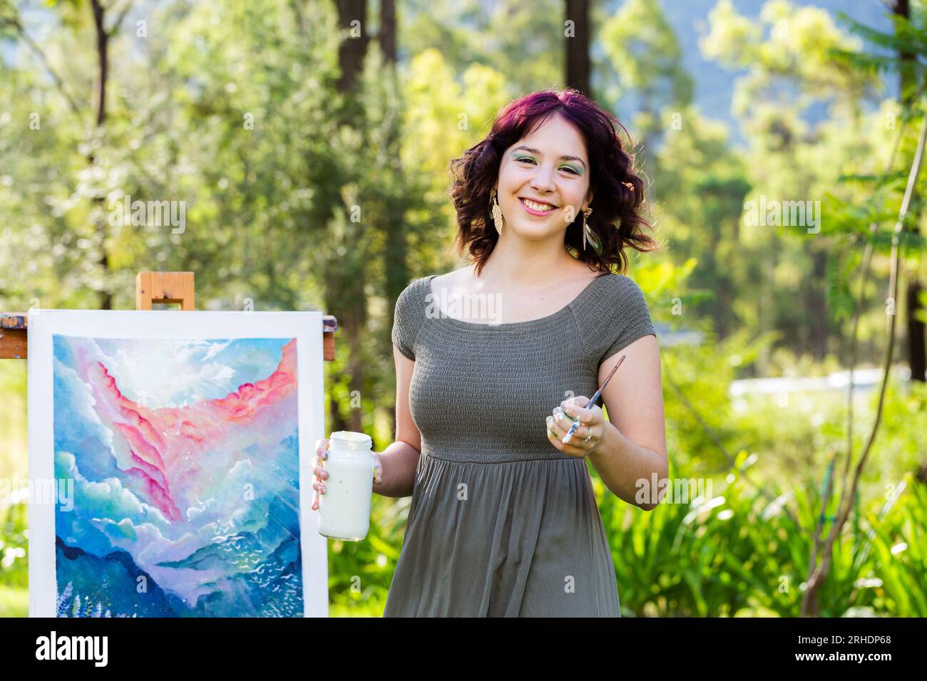 Felice giovane artista australiano che tiene un vaso di vernice bianca e un pennello accanto alle opere d'arte Foto Stock