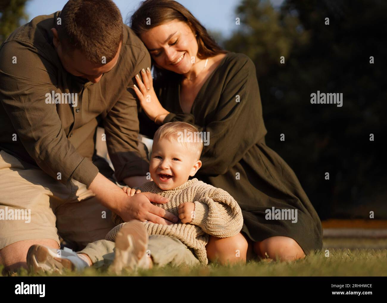 famiglia felice seduta sull'erba nel parco. Mamma che mette la testa sulla spalla del marito. bambino che ride felicemente. Foto di alta qualità Foto Stock