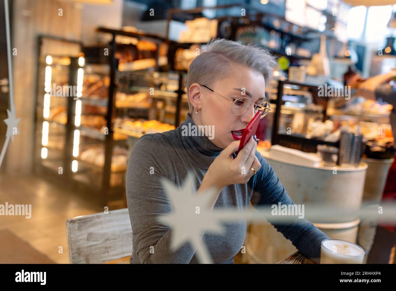 giovane donna bionda caucasica che registra un messaggio audio con lo smartphone seduto al bar. Visualizza tramite vetro, immagine con messa a fuoco selettiva Foto Stock