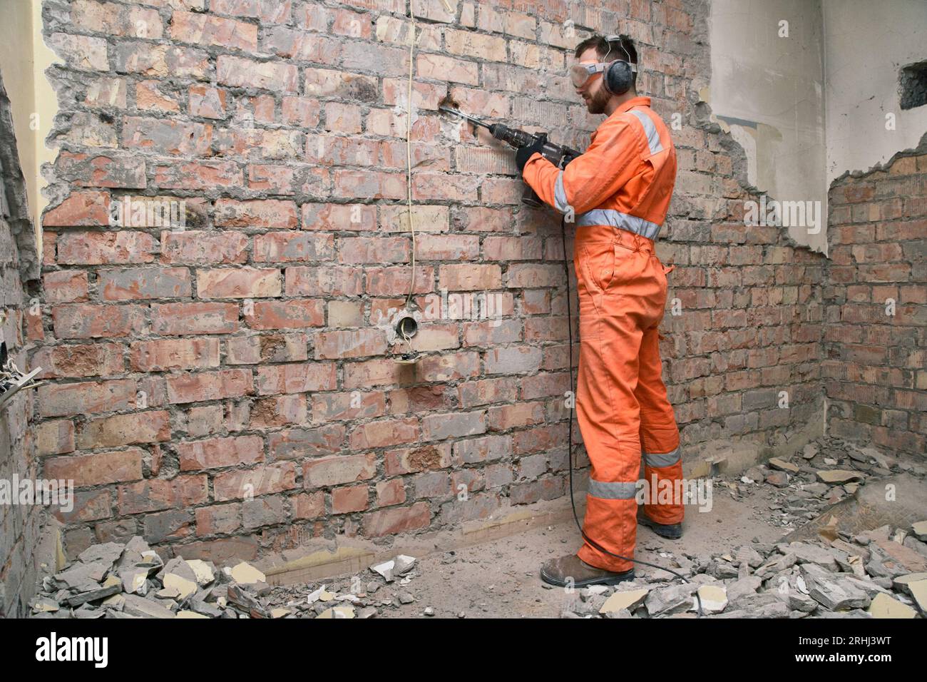 Lavoratore in piedi che rimuove il vecchio intonaco dal muro di mattoni rossi con un martello perforatore, indossando guanti, stivali, maschere, cuffie antirumore e tute arancioni. Foto Stock