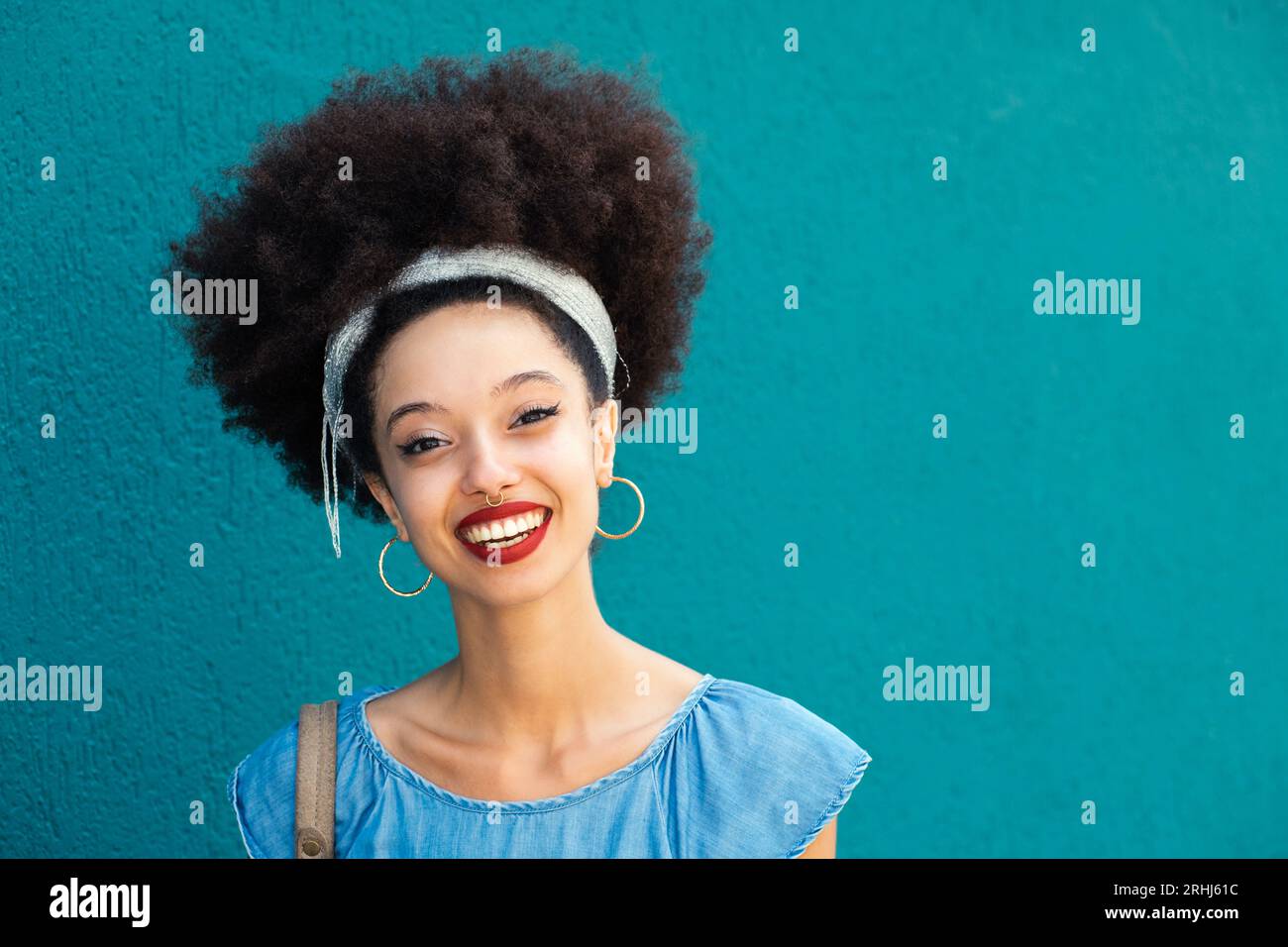 Ritratto di una giovane donna afro-americana ottimista con acconciatura afro e orecchini con trucco sorridente e guardando la fotocamera contro lo sfondo blu Foto Stock