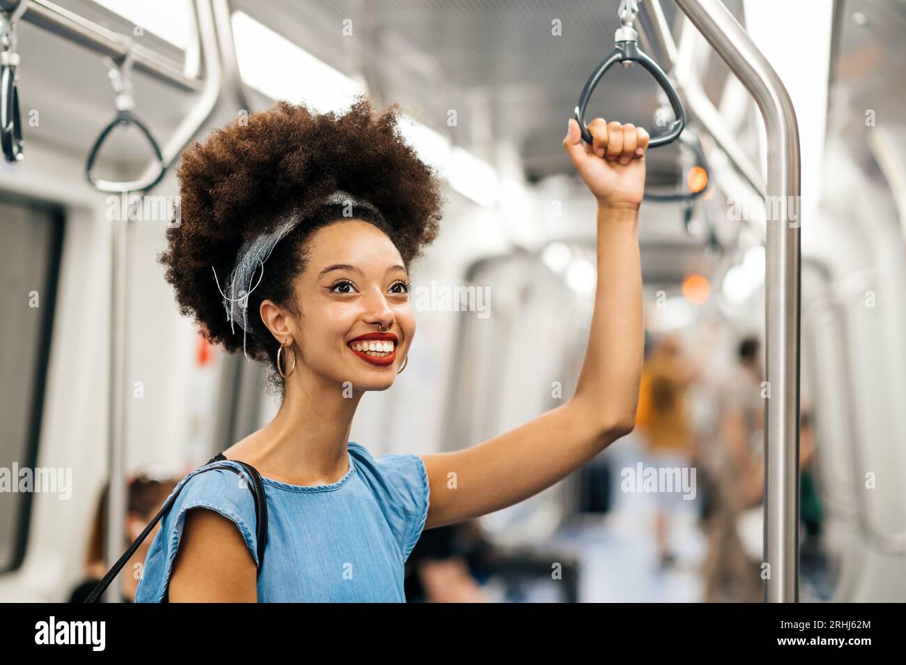 Giovane donna marocchina moderna e positiva con fascia per capelli afro che sorride e guarda lontano mentre si tiene la maniglia in piedi durante il viaggio Foto Stock