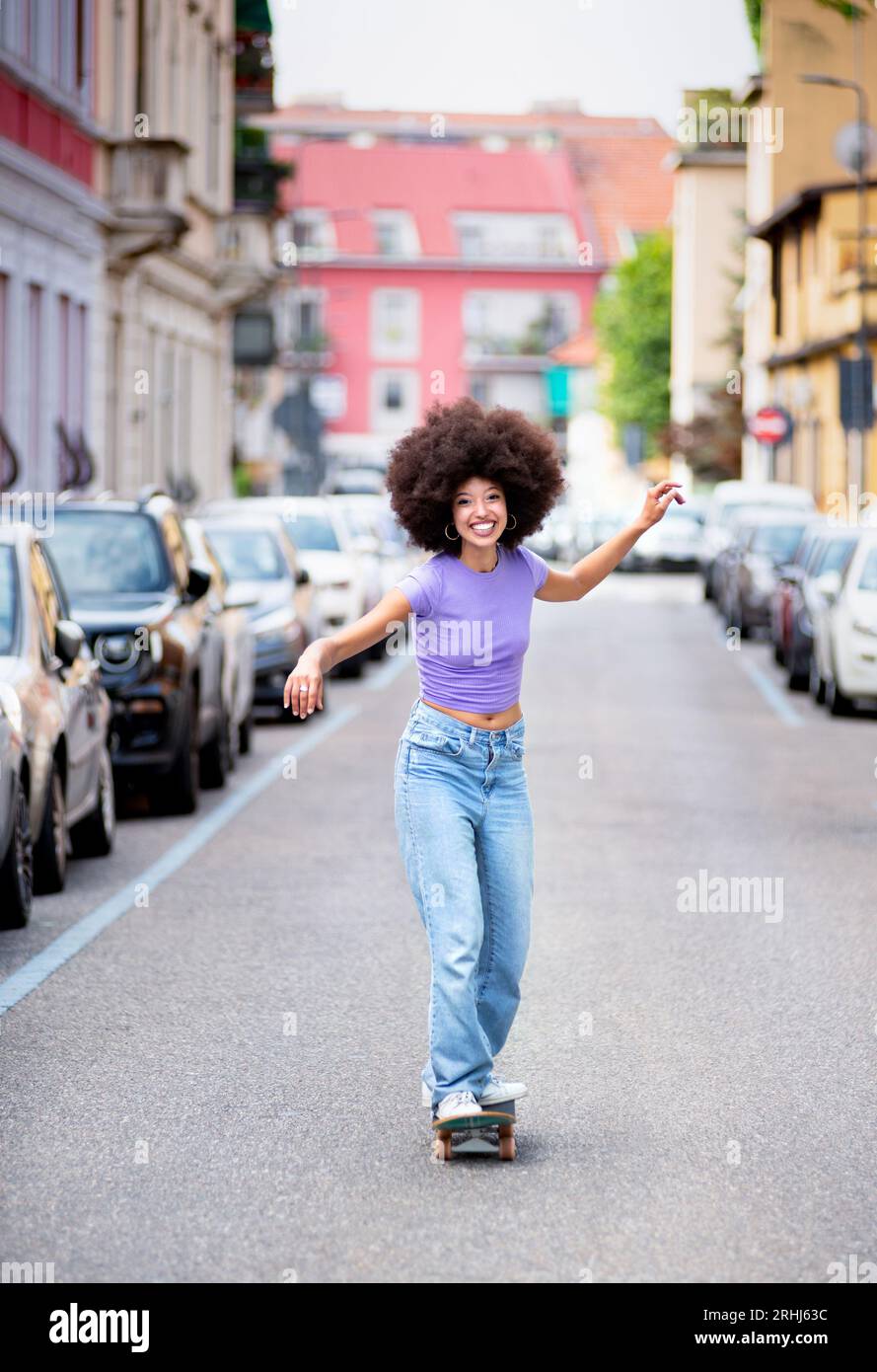 Corpo completo di allegra donna afroamericana in abiti casual con capelli afro che guardano la macchina fotografica mentre si guida sullo skateboard su strada asfaltata vicino al parcheggio c Foto Stock