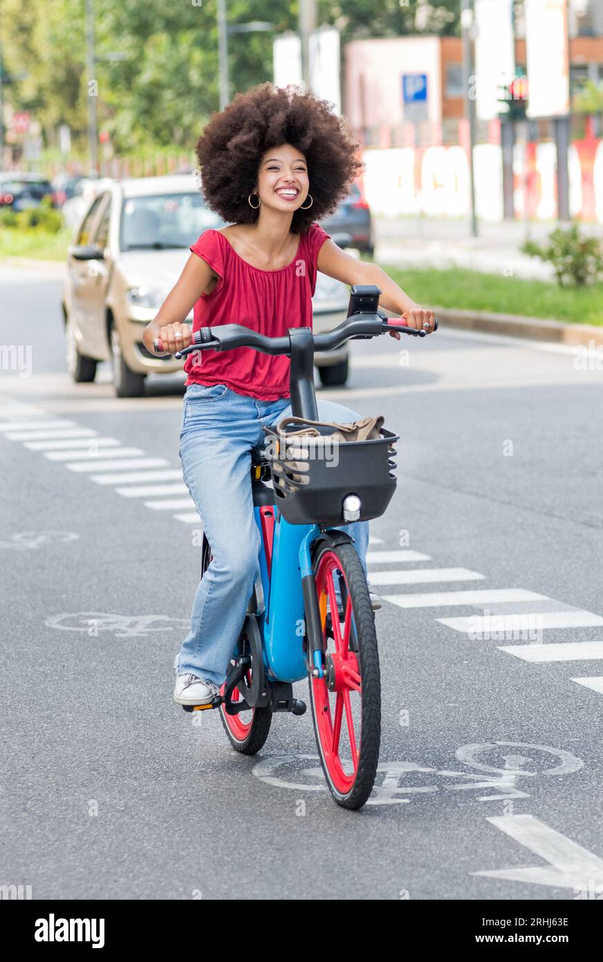 Giovane donna marocchina positiva con i capelli afro che guarda lontano mentre si guida in bicicletta elettrica con cesto su strada asfaltata alla luce del sole contro sfocato Foto Stock