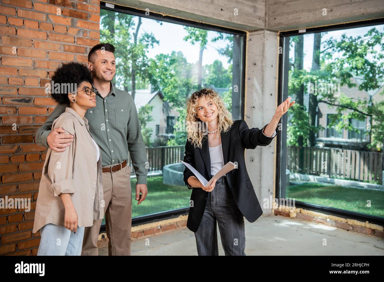 un broker immobiliare sorridente che punta con la mano mentre mostra nuove abitazioni a coppie multietniche Foto Stock