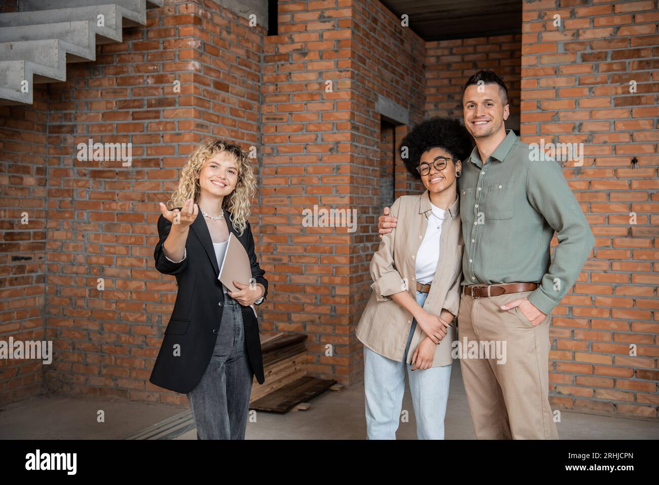 un broker immobiliare sorridente che punta con mano e mostra nuove abitazioni a coppie multiculturali Foto Stock
