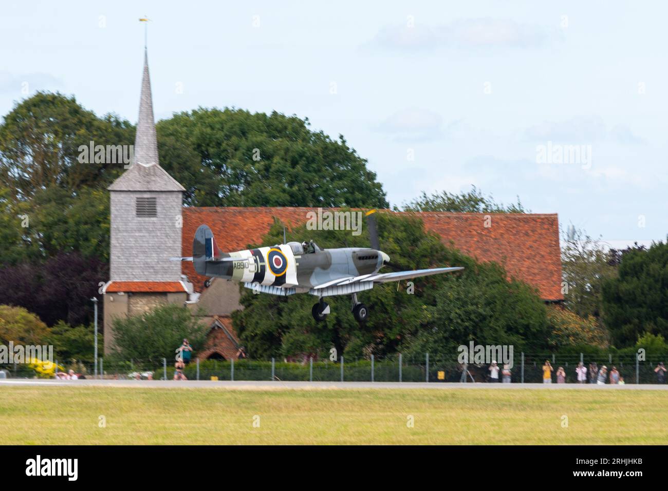 London Southend Airport, Essex, Regno Unito. 17 agosto 2023. Gli aerei d’epoca della RAF Battle of Britain Memorial Flight stanno utilizzando l’aeroporto civile relativamente tranquillo dell’Essex per operare per gli spettacoli aerei sul mare di questo fine settimana a Eastbourne e Folkestone. Il combattente Spitfire è raffigurato atterrando, passando per St Laurence e la Chiesa di Ognissanti sul perimetro dell'aeroporto. La gente è venuta a vederli arrivare Foto Stock