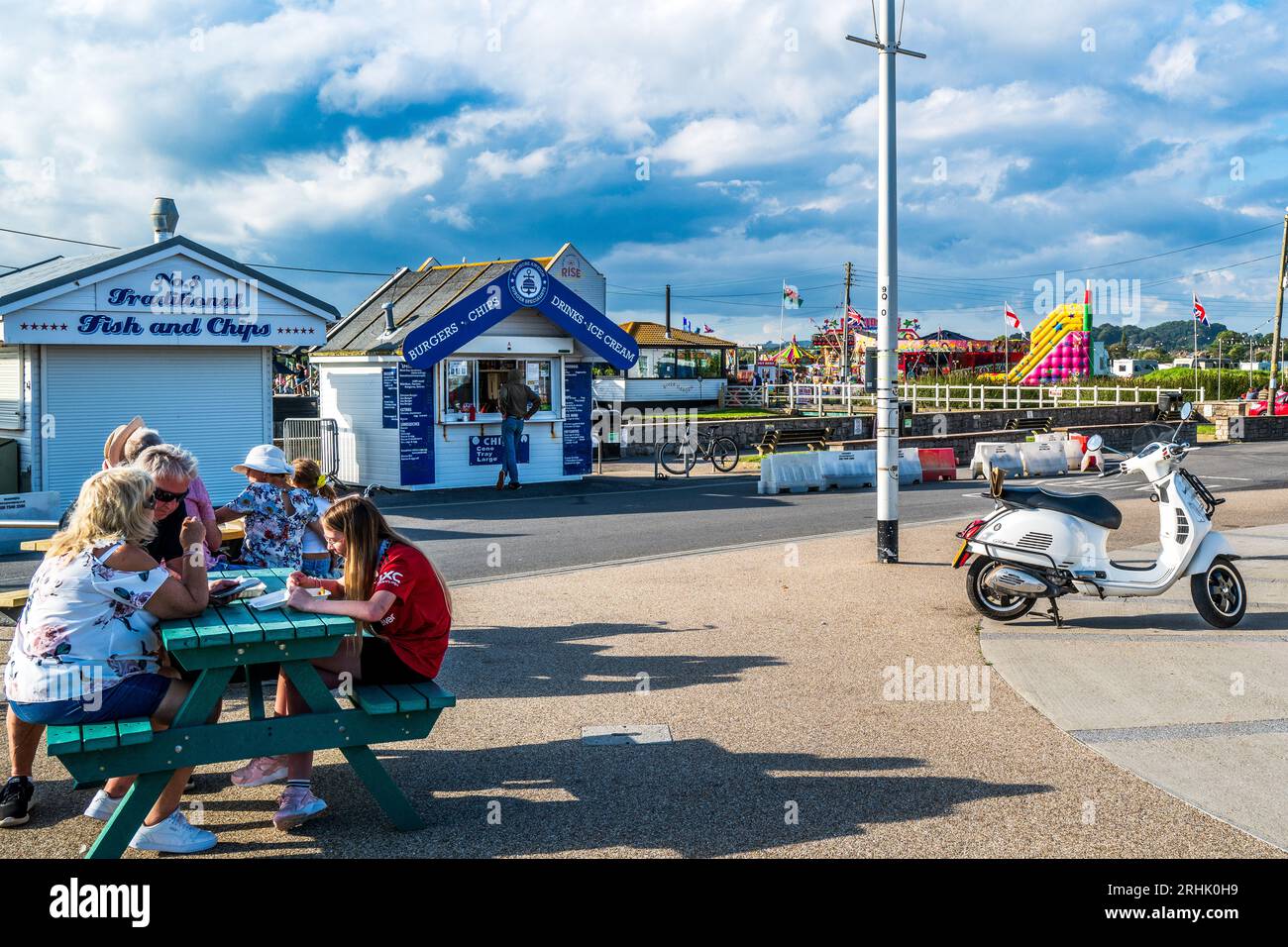 Turisti fuori da una baracca fish and chip, West Bay, Dorset, Regno Unito. Classica scena balneare britannica, scooter lucido, fish & chips, nuvole e luna Park Foto Stock