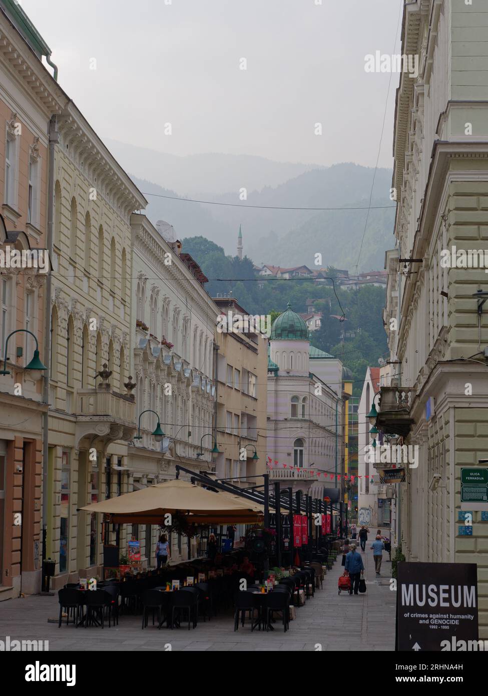 Pittoresca strada costeggiata da un bar con minareto sulle colline nebbiose alle spalle in una mattinata estiva a Sarajevo, Bosnia ed Erzegovina, 18 agosto 2023. Foto Stock