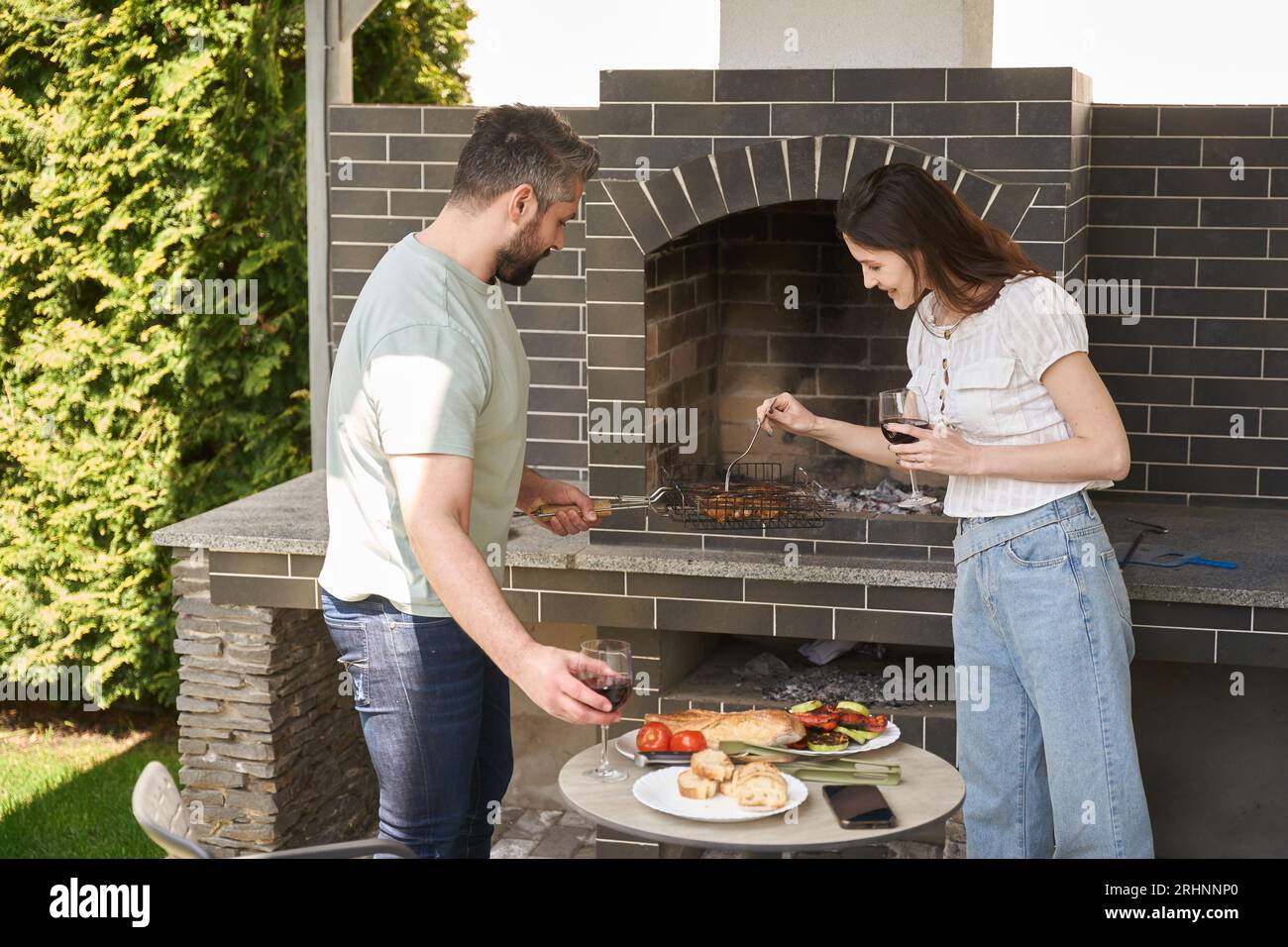 Gli sposi cucinano il cibo in un grande forno nel cortile sul retro, le persone indossano abiti comodi e informali Foto Stock