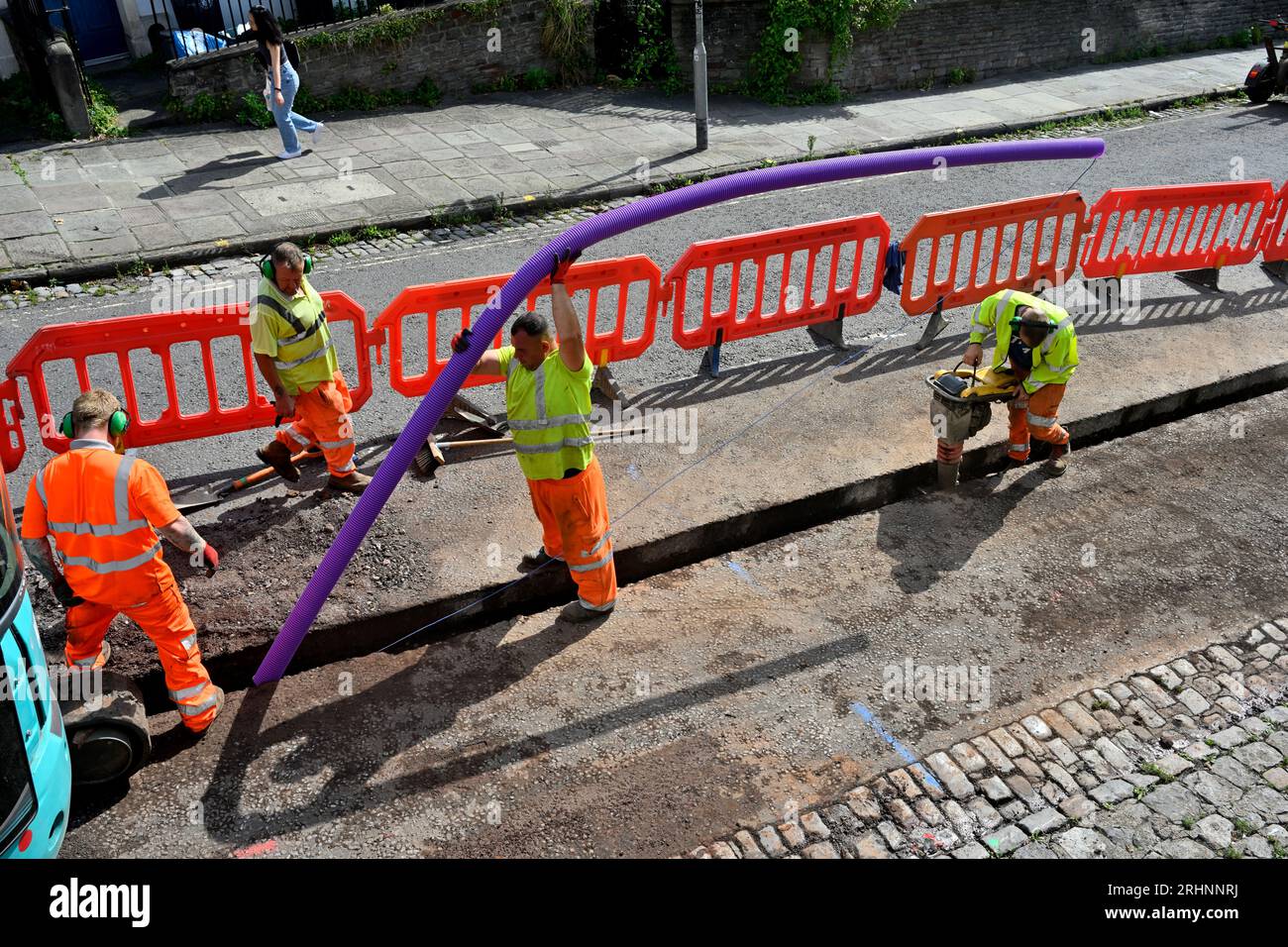 Operai che installano condotti per cavi in fibra ottica in trincea tagliata su strada con vibrante che compattano il fondo della trincea prima che il condotto sia in posizione, Regno Unito Foto Stock