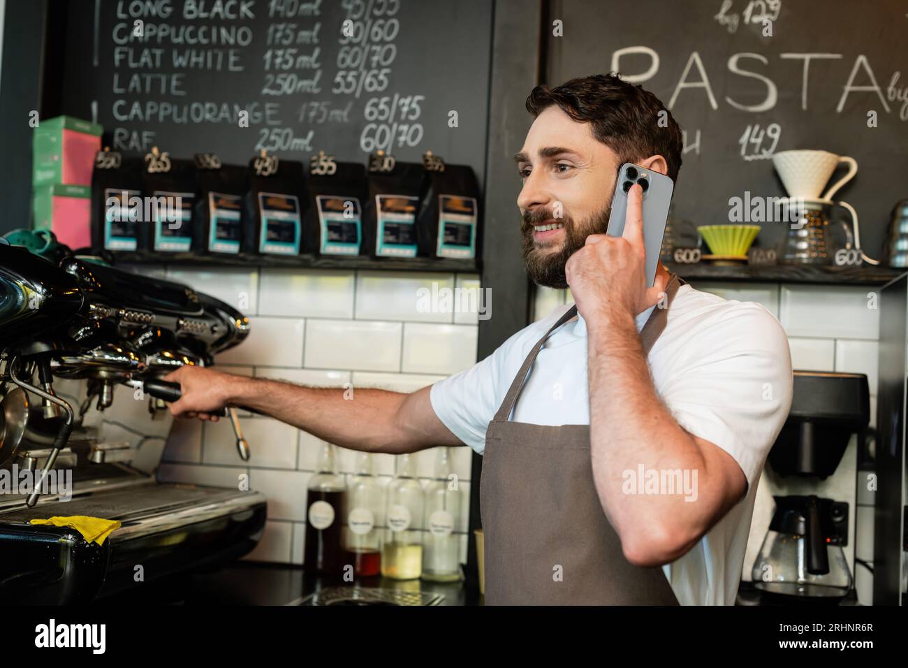 barista sorridente in grembiule che parla sullo smartphone mentre lavora vicino alla macchina del caffè nel bar Foto Stock