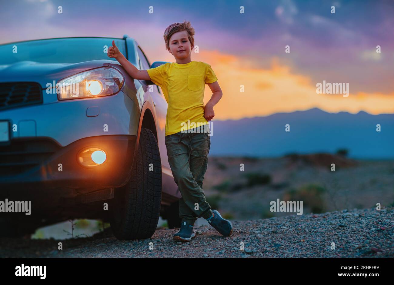 Ragazzo di successo in piedi accanto all'auto e mostrando i pollici al crepuscolo sullo sfondo delle montagne Foto Stock