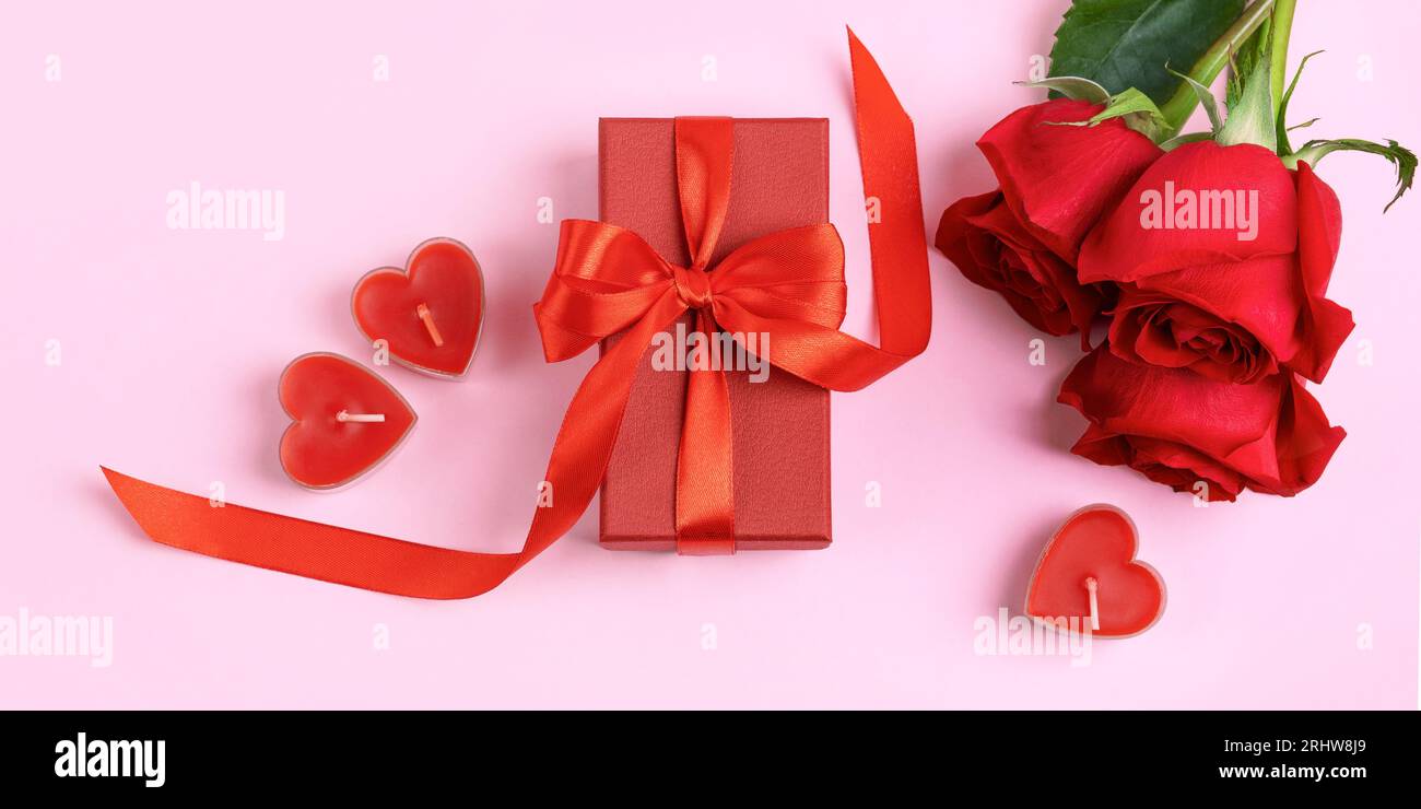 Rose rosse e confezione regalo rossa con fiocco di raso rosso e candele a forma di cuore. Sfondo romantico con spazio per il testo. Buon saluto auto Foto Stock
