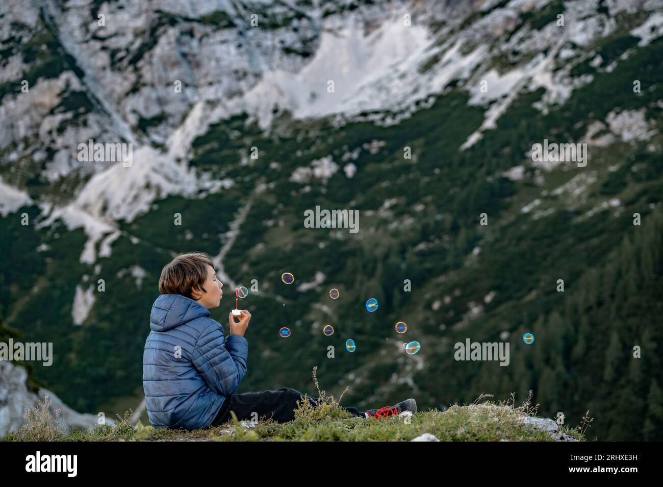 Adolescente seduto su una roccia coperta di muschio in alta montagna, prendendosi una pausa dalle escursioni e soffiando casualmente bolle di sapone. Bellezza e semplicità di li Foto Stock