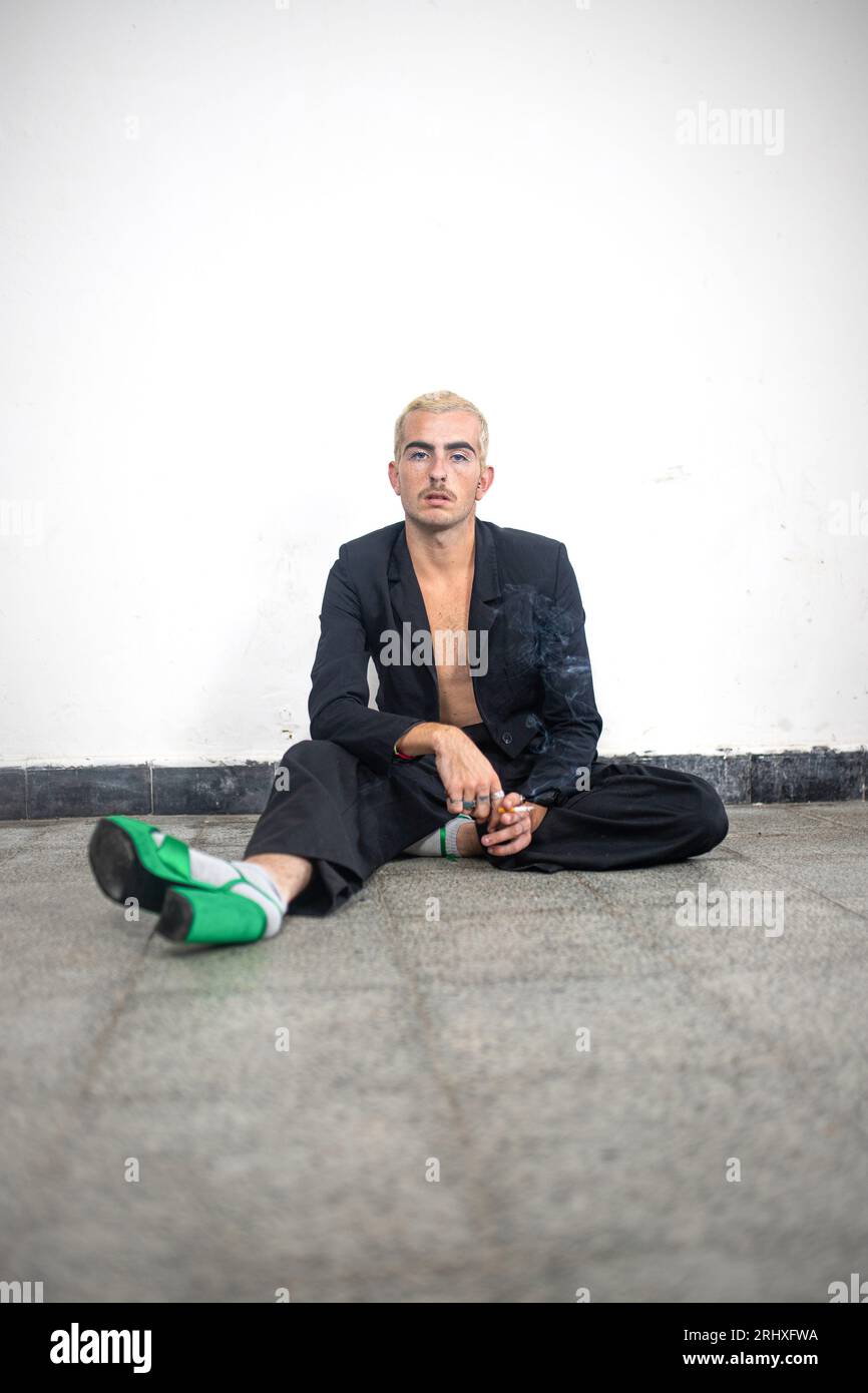 Uomo sicuro con tacco alto che guarda la fotocamera seduto da solo con la gamba allungata sul pavimento in marmo di una stanza vuota contro la parete bianca alla luce del giorno Foto Stock