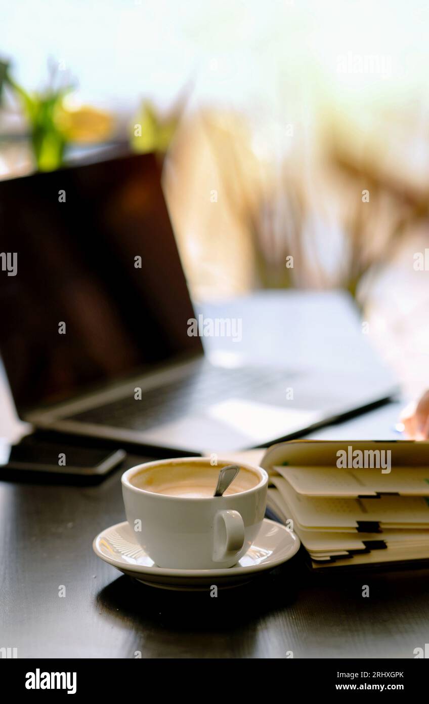Messa a fuoco selettiva di caffè cremoso in tazza con cucchiaio e notebook sul tavolo con sfondo sfocato di laptop e telefono aperti al bar alla luce del giorno Foto Stock