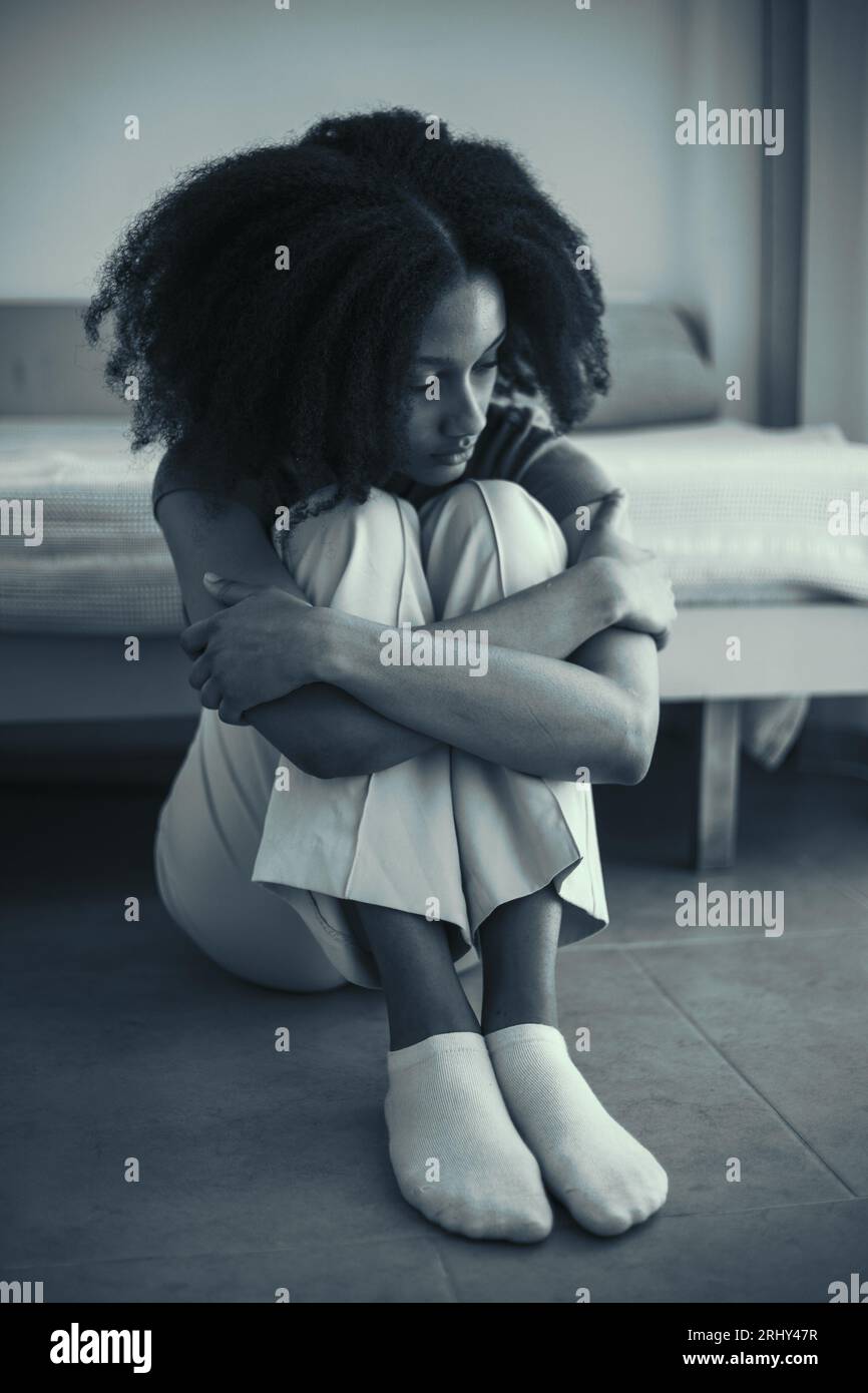 Una graziosa ragazza nera in stress emotivo si siede da sola sul pavimento a casa. Giovane adolescente preoccupato per i problemi a scuola e il bullismo. Immagine in bianco e nero Foto Stock