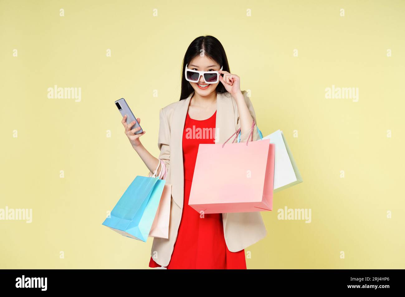 Giovane donna che tiene le borse della spesa mentre usa il telefono cellulare isolato su sfondo giallo. Foto Stock