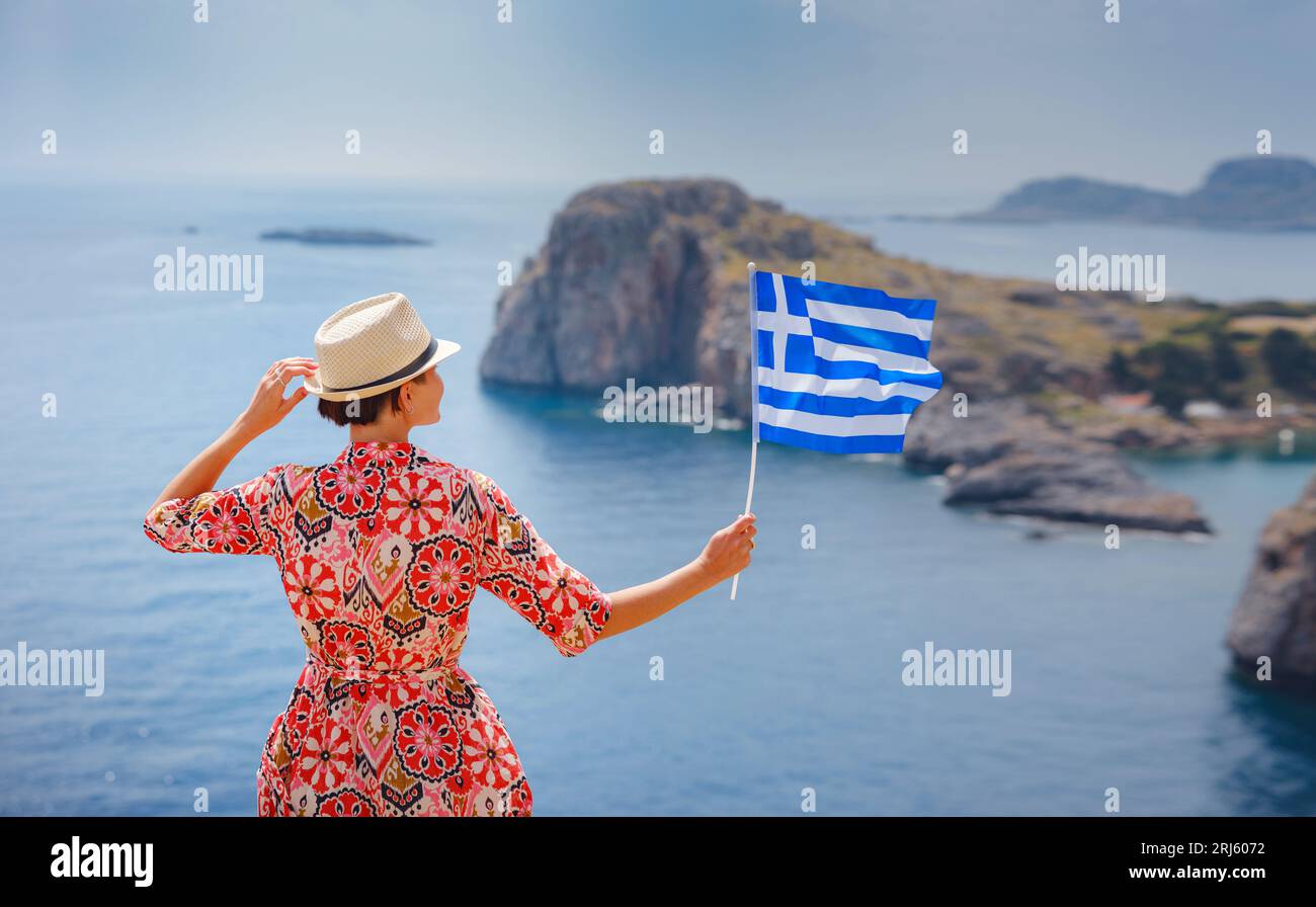 Bella e felice donna che si gode il sole alle isole greche. Viaggio in Grecia, isole del Mediterraneo fuori stagione turistica. Giovane donna viaggiatrice con bandiera greca nazionale che gode di una vista sul mare Foto Stock