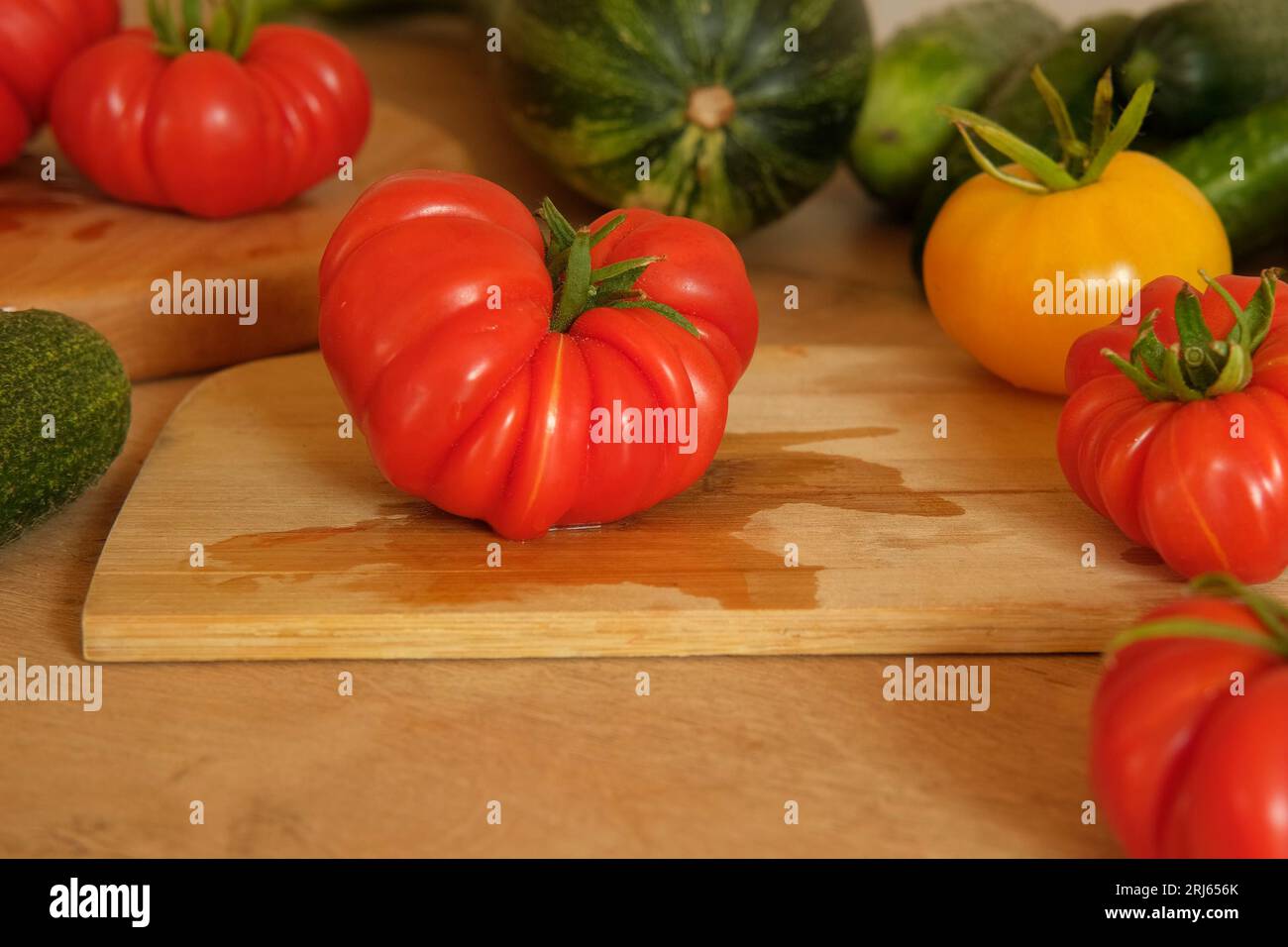 Un pomodoro grande maturo fresco su un tagliere di legno. Molte verdure crude di fondo. Pomodori, cetrioli. Fornello a casa che fa un pasto vegetariano sano. Foto Stock