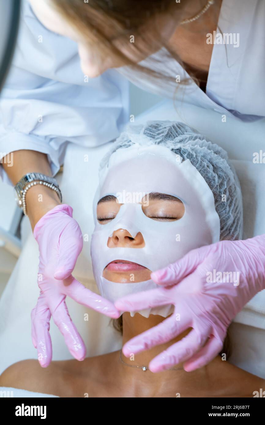 Cosmetologo che indossa guanti di gomma applicazione maschera foglio sul viso della donna. Foto Stock