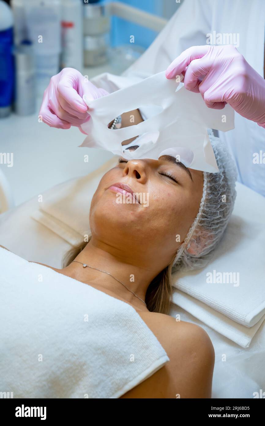 Estetista rimozione foglio maschera sul viso femminile per reidratare la pelle del viso, anti invecchiamento procedura cosmetica. Foto Stock