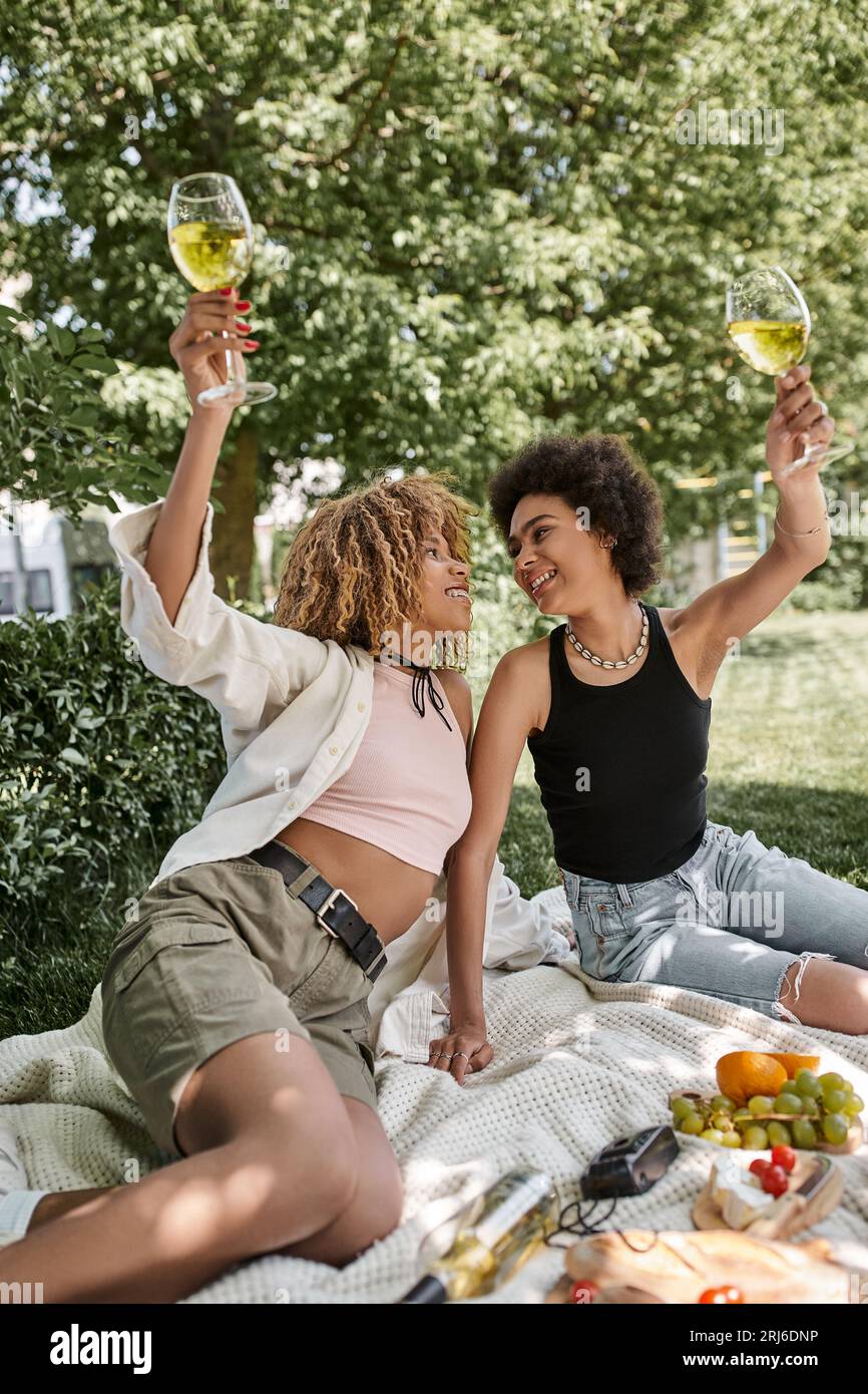 eccitata donna afroamericana che brinda con un bicchiere di vino vicino alla fidanzata, divertimento, picnic nel parco Foto Stock