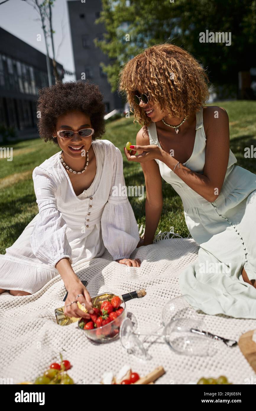 gioiose amiche afroamericane che mangiano fragole e uva fresche durante il picnic estivo Foto Stock