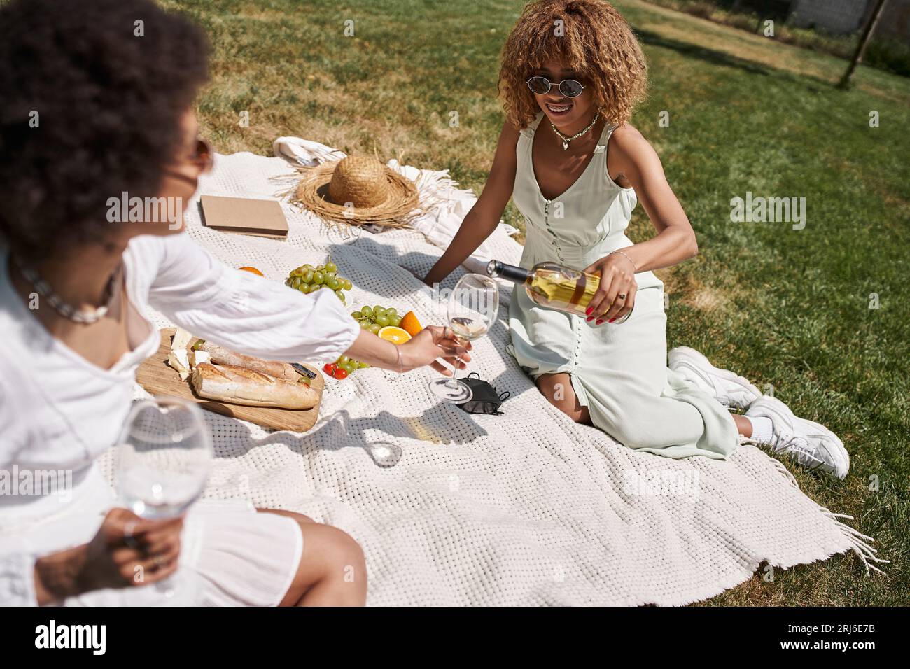 donna afroamericana che versa vino nel bicchiere della fidanzata durante il picnic estivo nel parco Foto Stock