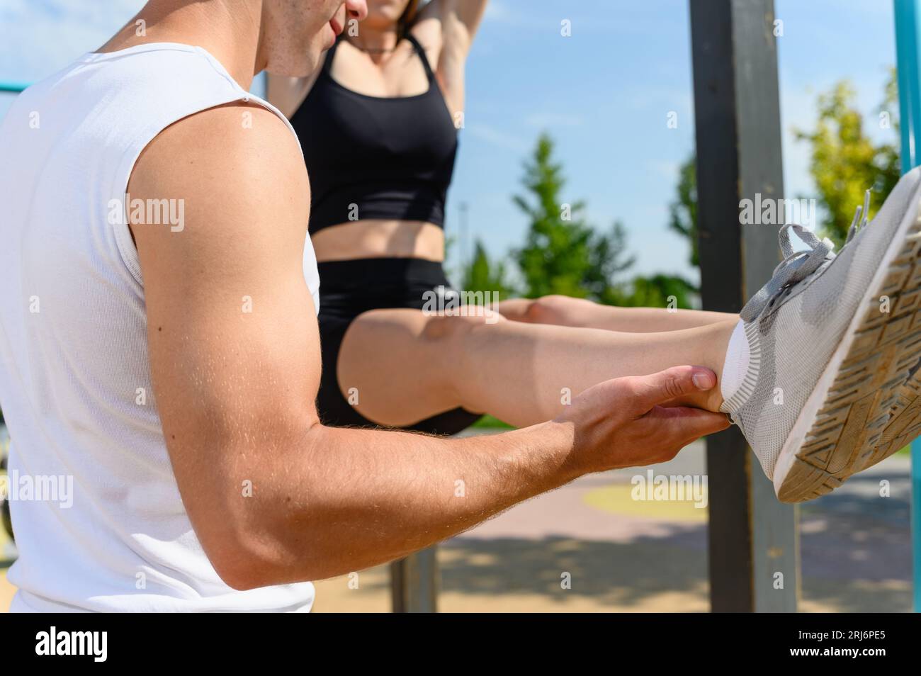 Le donne stanno facendo l'esercizio a L-sit e appese al bar orizzontale su un terreno sportivo di strada per un allenamento. L'uomo le tiene le gambe dritte e in bagno Foto Stock